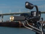 Sleek motorcycle handlebar with black grip and mirror, near water with bridge in background.