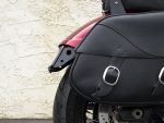 Motorcycle black leather saddlebag with buckle detail on a red and black motorcycle, displayed against a textured wall.