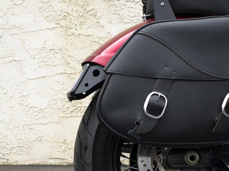 Motorcycle black leather saddlebag with buckle detail on a red and black motorcycle, displayed against a textured wall.