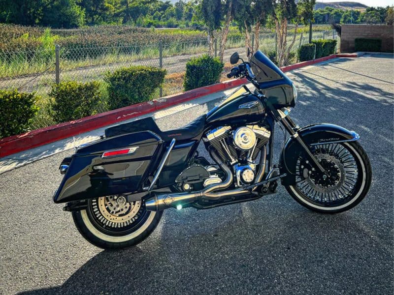 Black cruiser motorcycle parked on asphalt with custom wheels and leather seat in an outdoor setting.