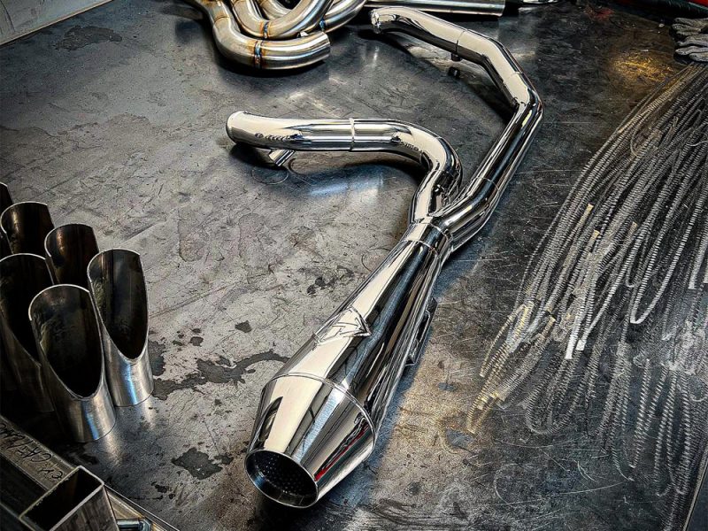 Shiny chrome motorcycle exhaust pipe with custom fabrication on a workshop floor.