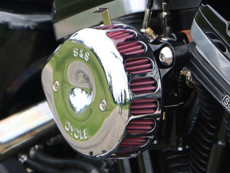 Chrome motorcycle engine component with purple air filters, reflecting outdoor scenery, branded S&S Cycle, high-performance parts for custom bikes.